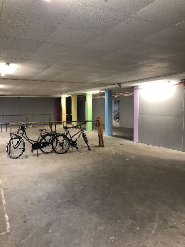 Das Bild zeigt einen Teil des Fahrradkellers nach der Streichaktion. Die Wände wurden in hellgrau und dunkelgrau gestrichen, die vorhandenen Säulen in den Farben des Regenbogens. Zudem sind im Vordergrund des Bildes zwei Fahrräder zu sehen. 